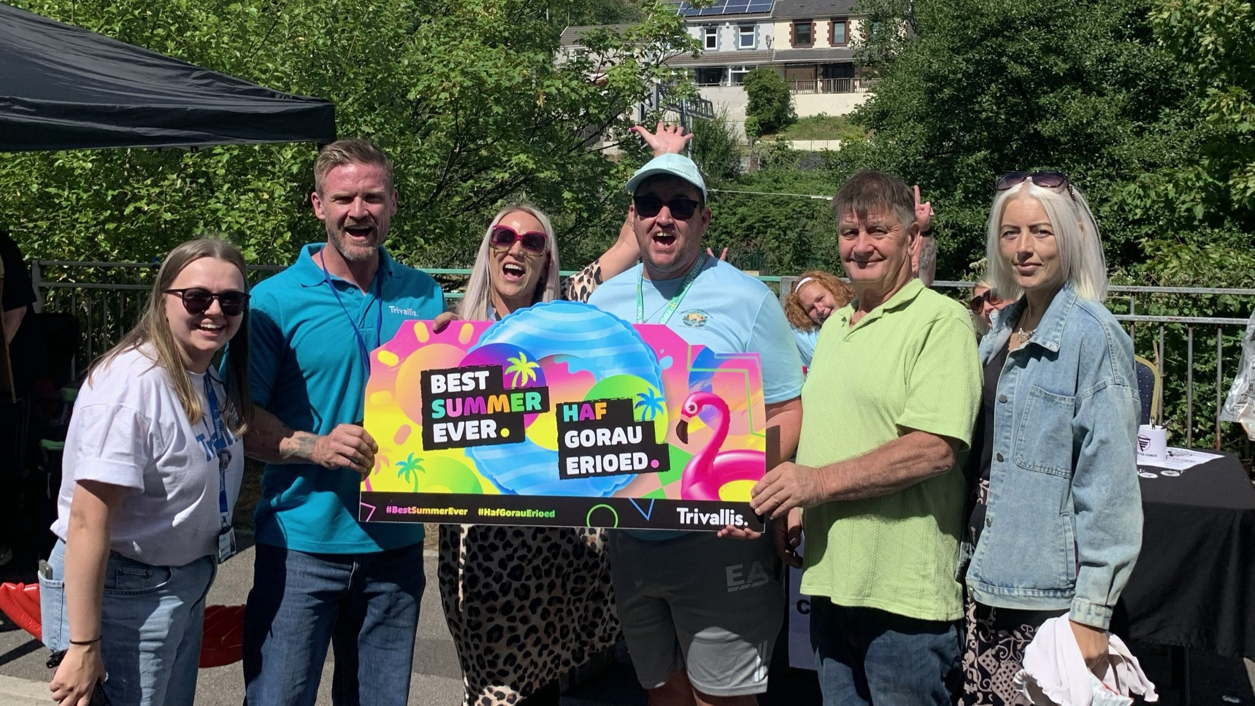 Trivallis Housing Landlord Wales Six people stand outside in the sun holding a sign that says “Best Summer Ever” and “Haf Gorau Erioed” in English and Welsh.