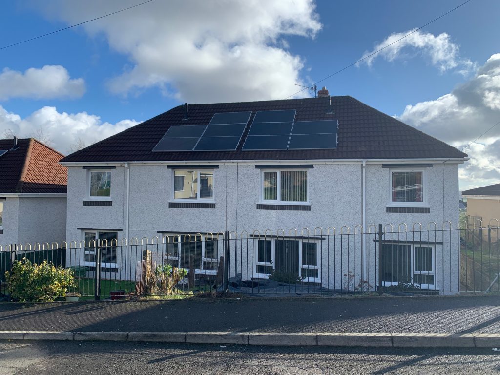 A two-story white pebbledash house with solar panels on the roof, black gates at the front, and a blue sky with scattered clouds above.