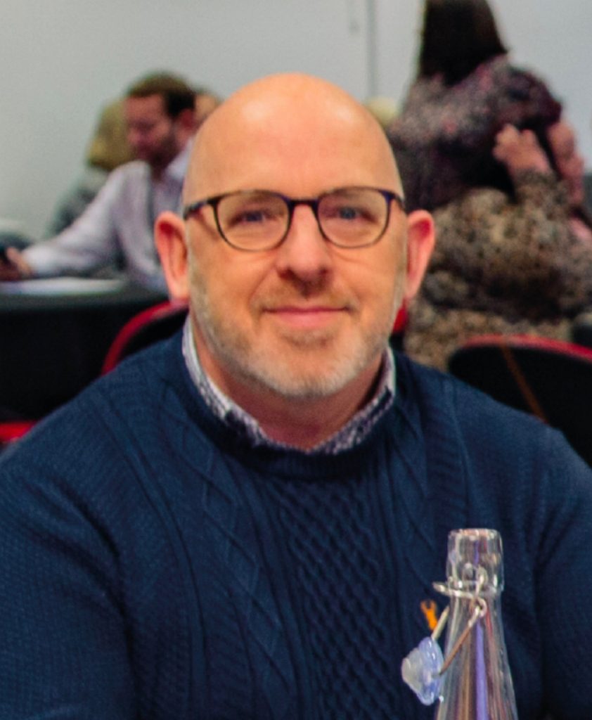 A bespectacled man with a short beard and bald head, wearing a blue sweater over a patterned shirt, smiling at the camera with people in the background.