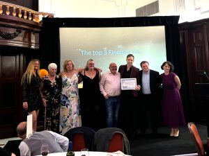 Trivallis Housing Landlord Wales A group of eight people stands together on a stage in front of a large screen displaying the text "The top 3 Finalists are...". One person in the group is holding a framed award. The background features ornate wooden paneling and a high ceiling.