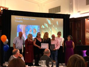 Trivallis Housing Landlord Wales A group of seven people are standing in a line on a stage, holding plaques. Behind them, a screen displays "Tenant Action Panel - Trivallis" along with an image of a person and some text. The setting appears to be an awards ceremony or formal event.