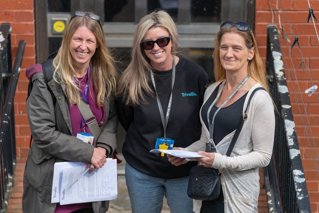 Trivallis Housing Landlord Wales Three women stand together, smiling for the camera. They are outdoors, in front of a brick building. Two women are holding documents, and one is wearing a "Trivallis" branded shirt. They are dressed in casual attire, and there's a staircase with railings visible beside them.