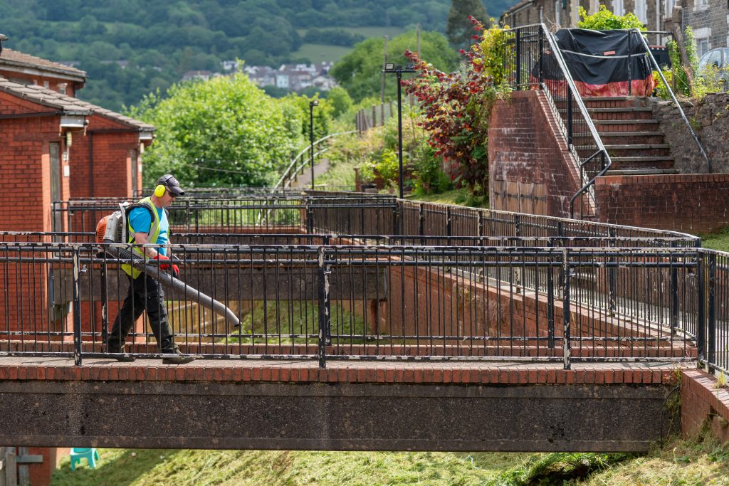 Trivallis Housing Landlord Wales A person wearing a high-visibility vest, ear protection, and a baseball cap is using a leaf blower on a footbridge. The surroundings include brick buildings, a grassy area below, and green hills in the background.