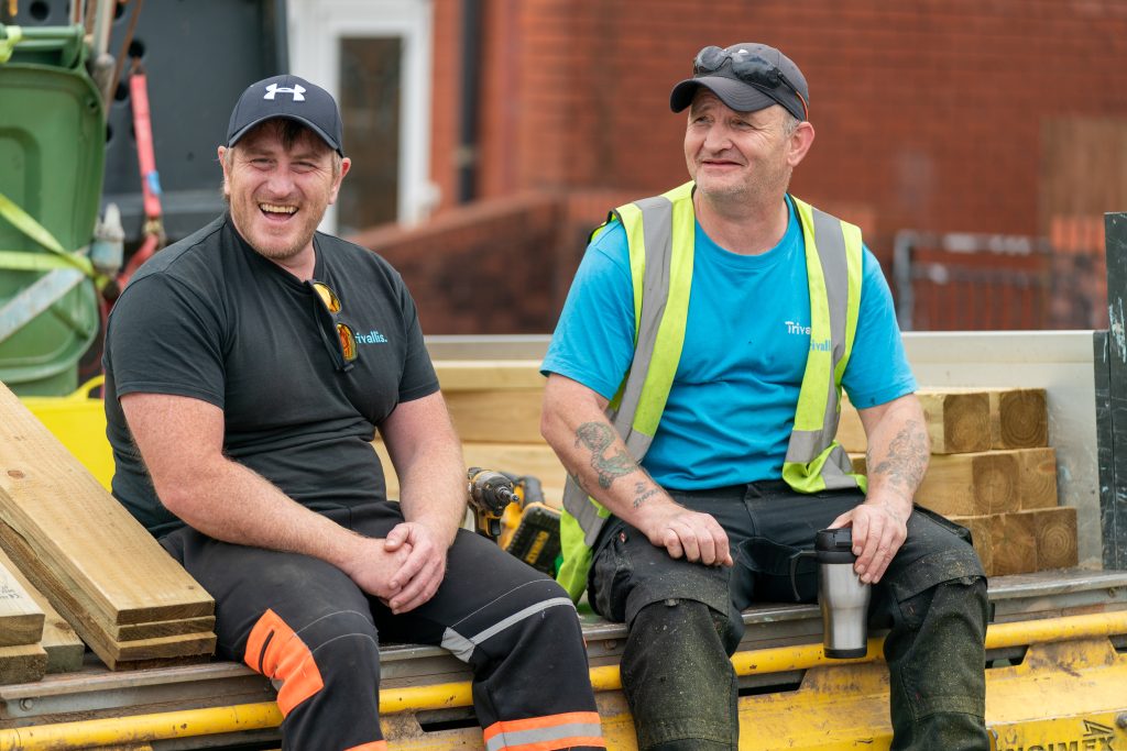 Trivallis Housing Landlord Wales Two construction workers sit on a flatbed truck among wooden planks and tools. Both are wearing casual hats, t-shirts, and work pants. One worker is smiling widely, while the other, wearing a high-visibility vest, calmly holds a travel mug. A brick building is in the background.